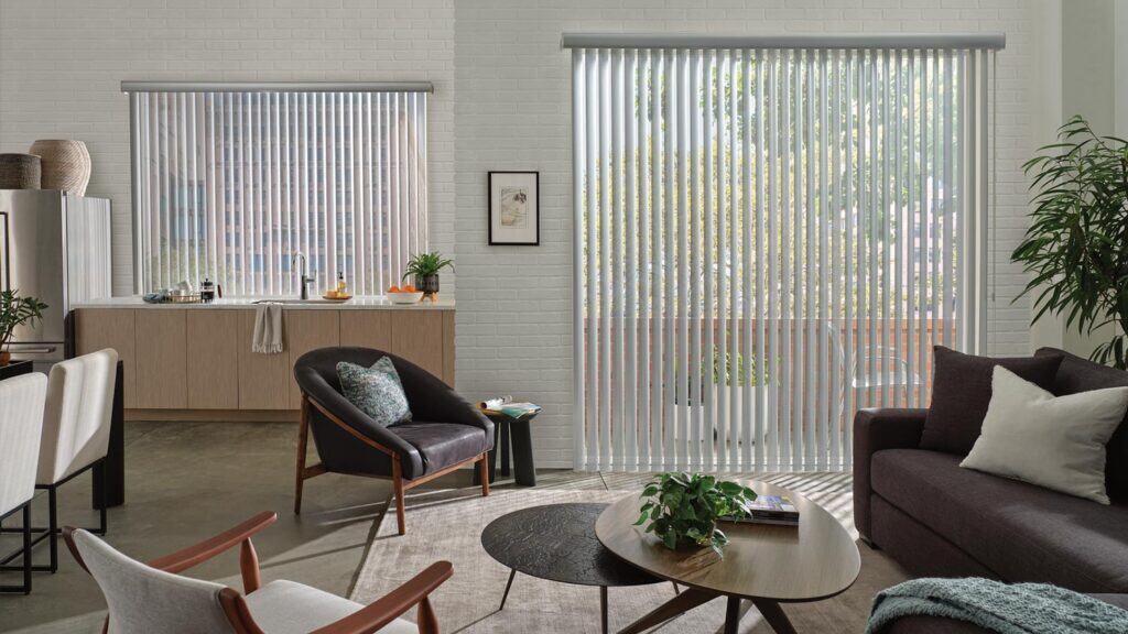 Bright living space featuring large windows with vertical blinds, cozy furniture, and indoor plants for a welcoming atmosphere, perfect for relaxed living and natural light.