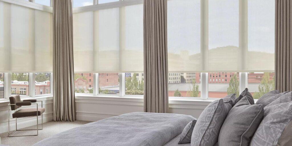 Bright bedroom featuring large windows, neutral curtains, and comfortable bedding overlooking downtown cityscape.